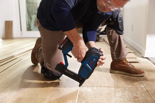 Una persona que lleva gafas de seguridad y rodilleras se arrodilla mientras usa una clavadora Bosch, 18V 2Ah, para instalar un suelo de madera.