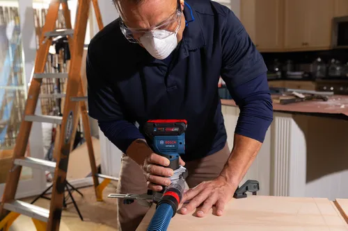 Persona utilizando una fresadora Bosch GKF18V-25 con una manguera de extracción de polvo, usando gafas de seguridad y una mascarilla.