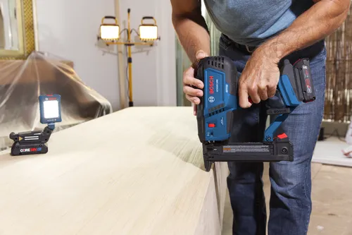 Clavadora Bosch GTH18V-18M, persona vestida con camisa azul y vaqueros, sosteniendo la clavadora, con "BOSCH" visible, cerca de una superficie de madera, con equipo de iluminación visible en el fondo.