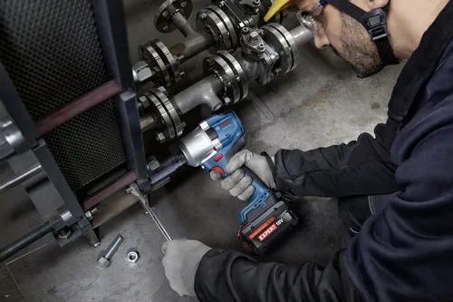 A worker using a Bosch GDS18V-780 18V Brushless Mid-Torque Impact Wrench to work on metal piping and flanges, wearing gloves and protective gear.