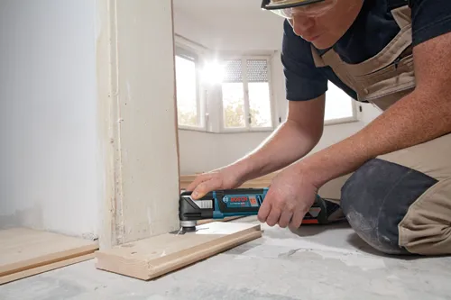 Persona utilizando multiherramienta de iones de litio Bosch, gafas de seguridad, en una habitación con suelo de madera.