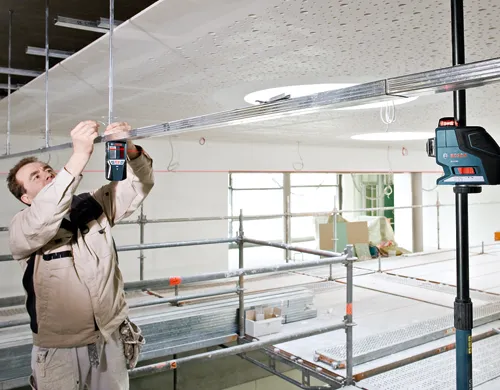 Un hombre con ropa de trabajo utiliza un nivel láser Bosch GLL 2-80 para instalar un techo suspendido.