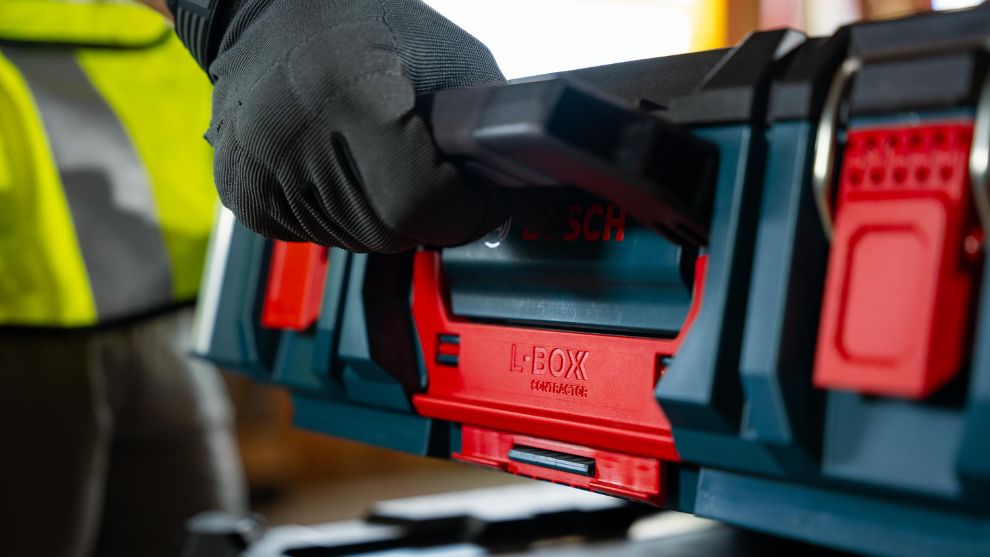 A gloved hand holds a blue Bosch Professional L-BOXX tool case, with red accents, in a bright workspace.